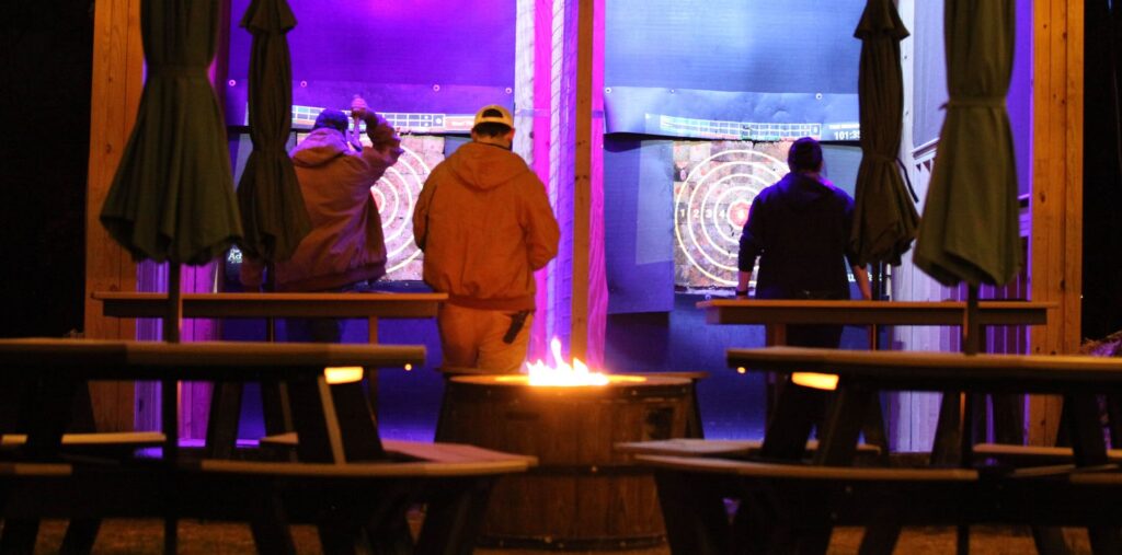 Adult axe throwing birthday party gathered at reserved tables and a fire pit. 